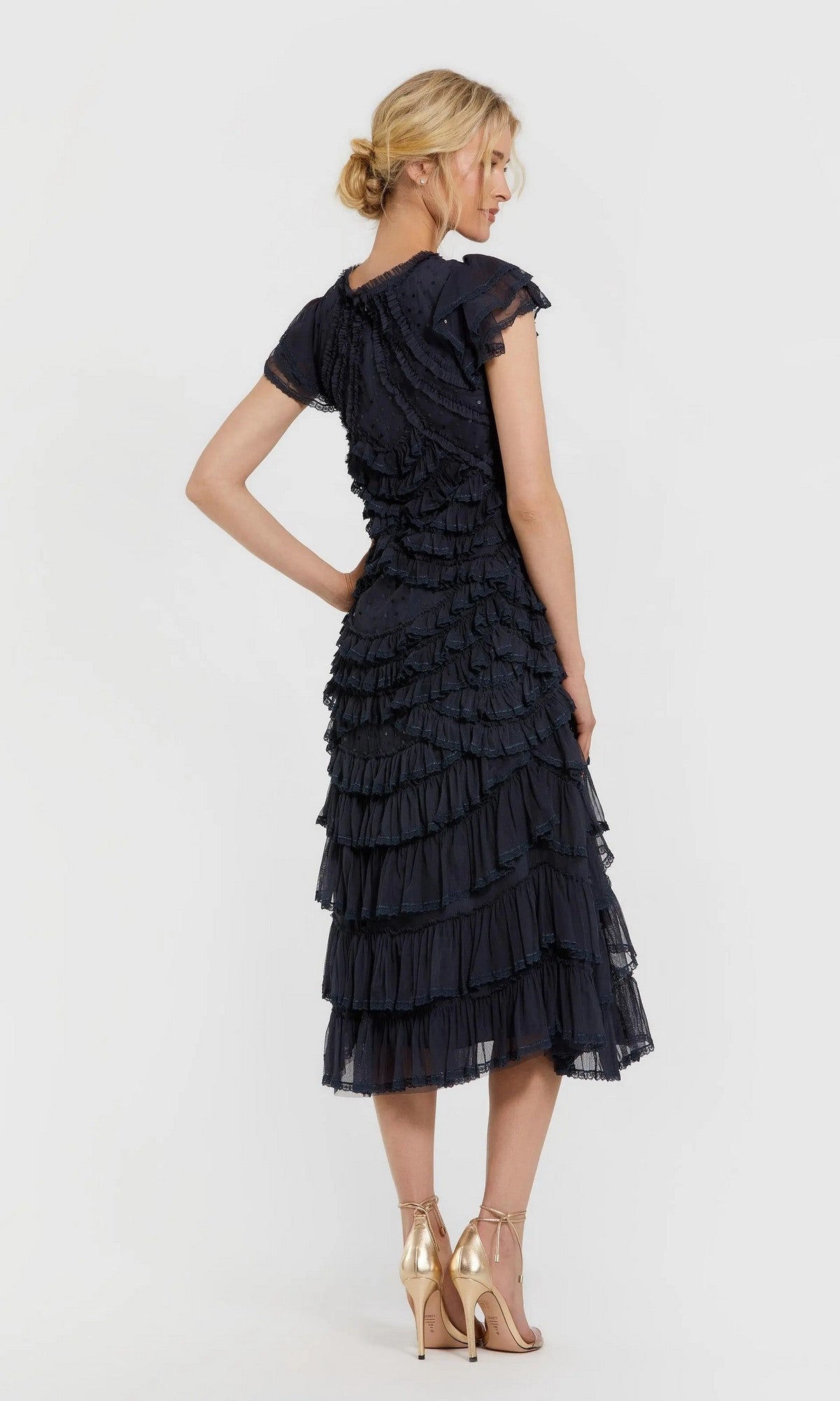 A woman stands in profile against a plain light background, wearing the Mac Duggal 8371 Midi Party Dress—navy blue with short sleeves, micro-ruffle detailing, and layered tiers—paired with gold strappy high-heeled sandals.
